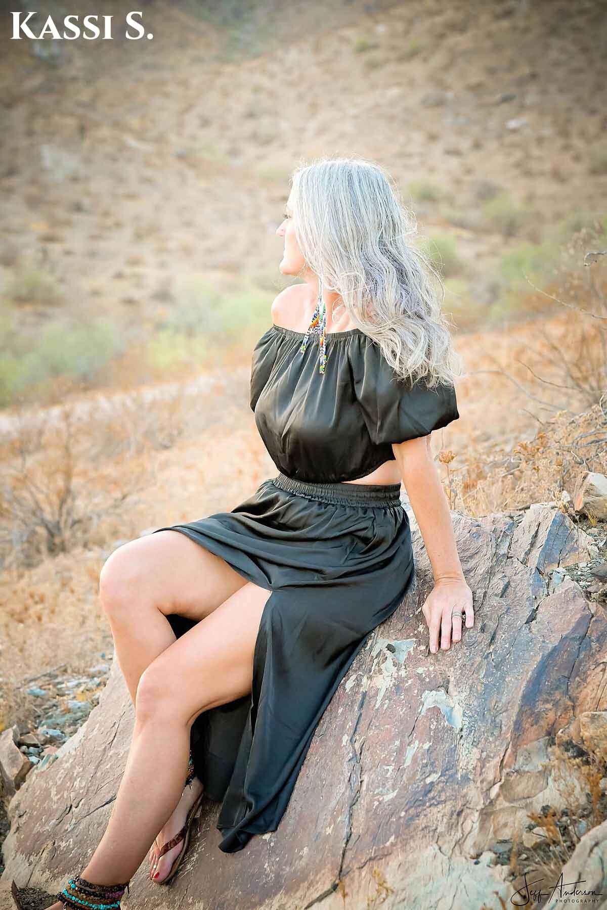 Woman with medium length gray hair sitting on a rock outside.